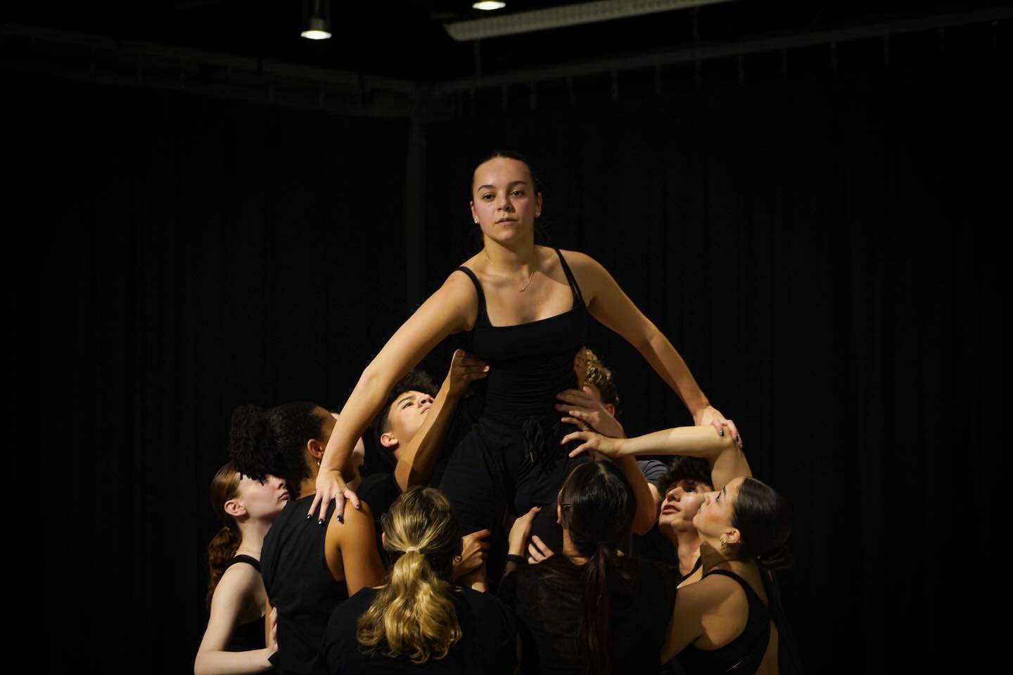 Ensemble of contemporary dancers in black costumes lift a female dancer at center stage under warm theatrical lighting.