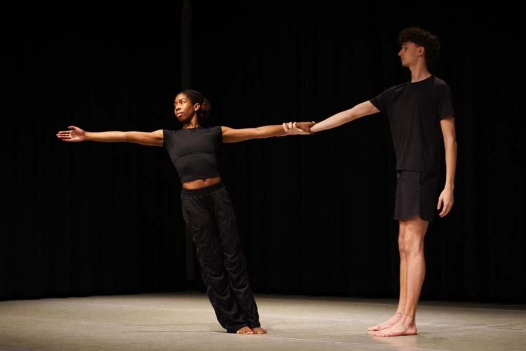 Two contemporary dancers onstage extend their arms while holding hands, balancing apart in a suspended duet.
