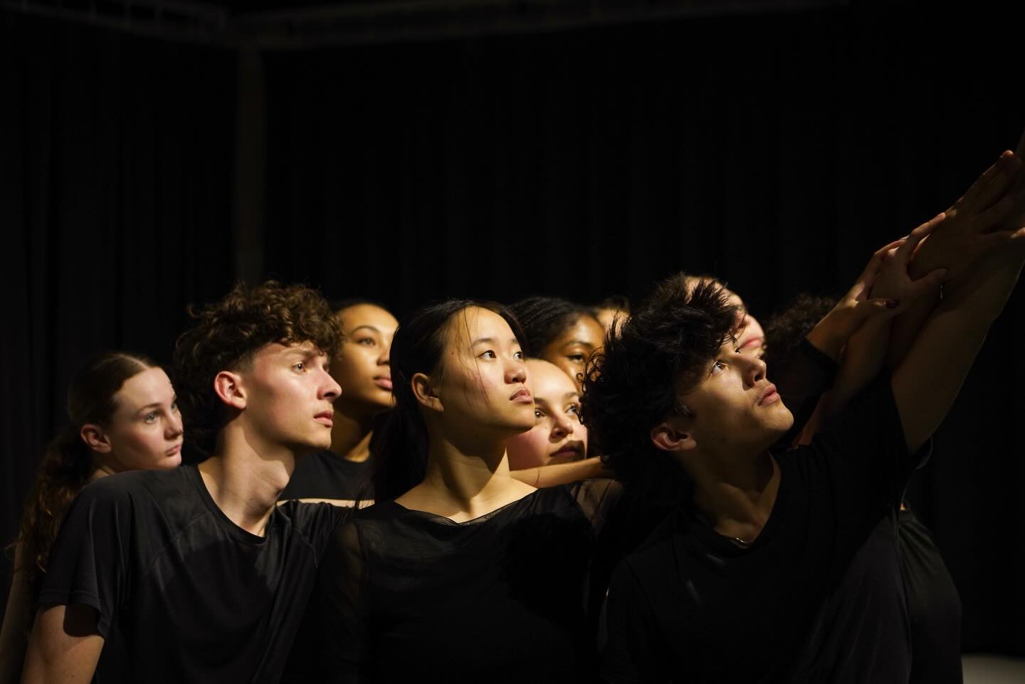 Close group formation of contemporary dancers gazing upward with arms raised under stage lighting.