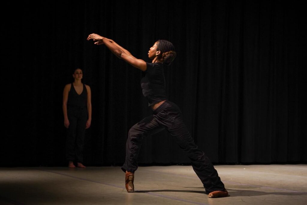 Contemporary dancer reaches forward in a deep lunge while another dancer stands in shadow behind.